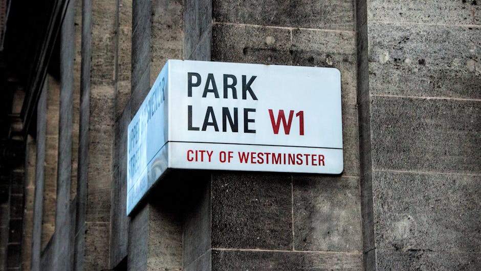 A white street sign with black and red text reading 'BUCKINGHAM PALACE ROAD SW1' and 'CITY OF WESTMINSTER' in red, mounted on two metal poles. The sign is positioned outside a building with a reflecti