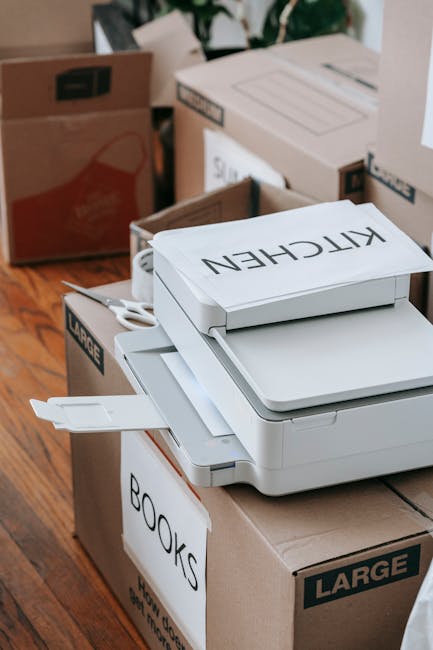 A close-up view of a stack of white paper photocopiers or printers with the brand name 'KITCHEN' printed on a paper label attached to the top device, situated on a brown cardboard box labeled 'BOOKS'