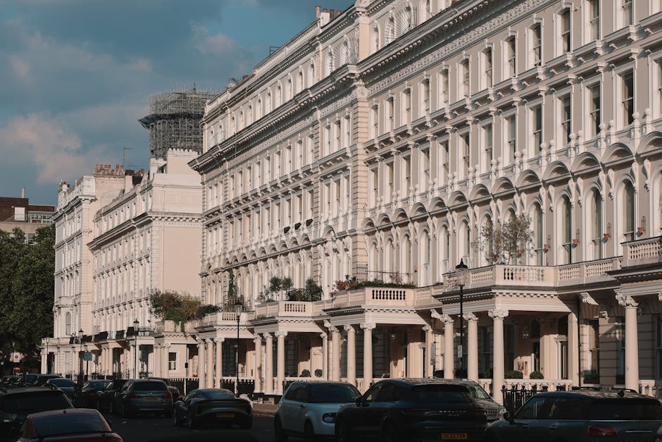 A row of elegant Victorian-style terraced houses with white facades and decorative black wrought iron balconies along a street, where several parked cars, including sedans and hatchbacks, line the pav